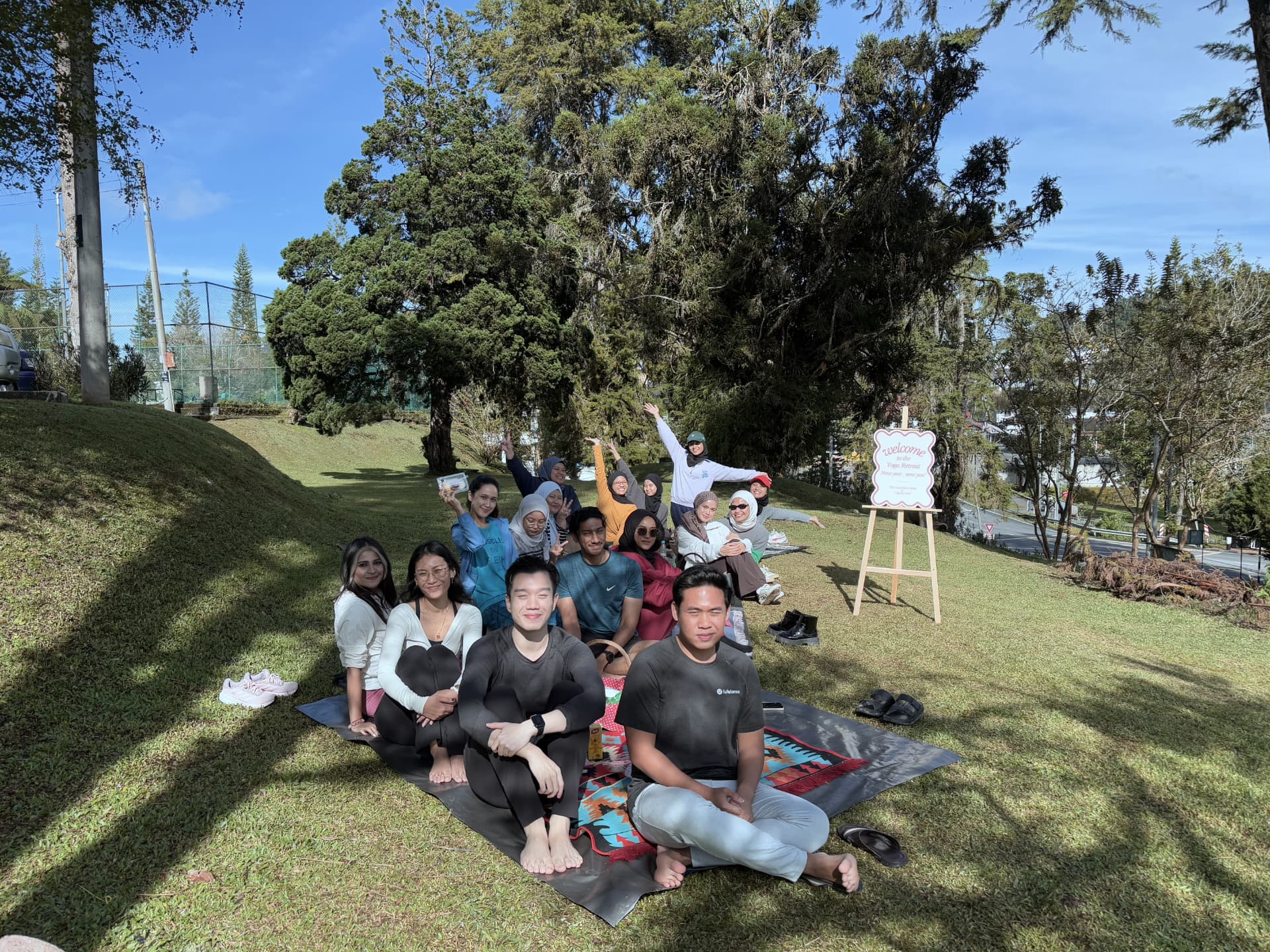 The full January 2025 Yoga Retreat group posed together on mats on a grassy hillside at The Cool Point Hotel, with the retreat welcome sign on an easel beside them